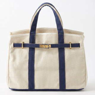 A canvas tote bag in natural beige with navy blue straps and trim, featuring a gold-tone metal buckle detail on the front, set against a plain white background.