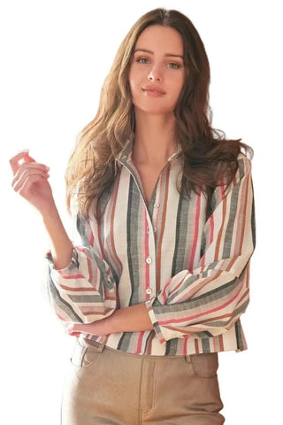 A woman with long wavy brown hair stands against a red brick wall wearing a Finley Crop Emmy Shirt with long-sleeve button-up shirt with vertical stripes in cream, red, and gray, paired with tan pants.