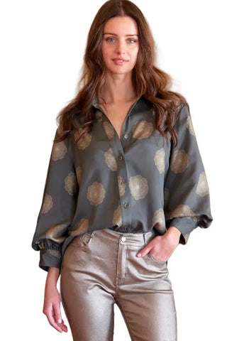 A woman stands in a doorway wearing a dark gray patterned button-up blouse with metallic gold circular motifs and shiny metallic silver cropped pants, paired with silver heeled sandals.
