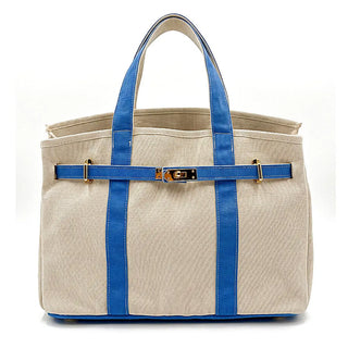 A structured canvas tote bag in beige with vibrant blue leather handles, side straps, and base, accented by a blue horizontal strap across the front with a gold-tone turn-lock clasp.