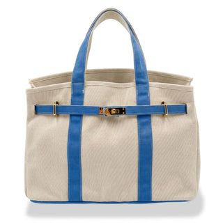 A unstructured boatkin tote bag in textured cream canvas with vibrant blue leather trim, double top handles, and a gold-tone turn-lock closure on a white background.