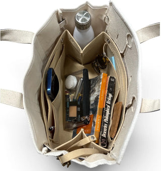 An open beige boatkin canvas tote bag with interior organizer pockets, shown from above on a white background. The bag contains various items including a white bottle, a blue compact, a black pen, and an orange book.