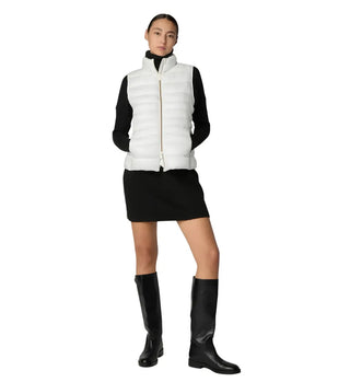 A model wearing a white puffer vest over a black long-sleeve top and black mini skirt, paired with black knee-high boots, on a solid white background.