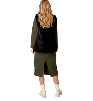 A woman shown from the back wearing a black faux-fur vest over an olive green long-sleeve top and matching olive green midi skirt, paired with off-white ankle boots, on a solid white background.