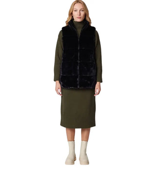 A model stands against a white studio background wearing an olive green knit long-sleeve dress with a black faux-fur vest layered over it, paired with off-white ankle boots.